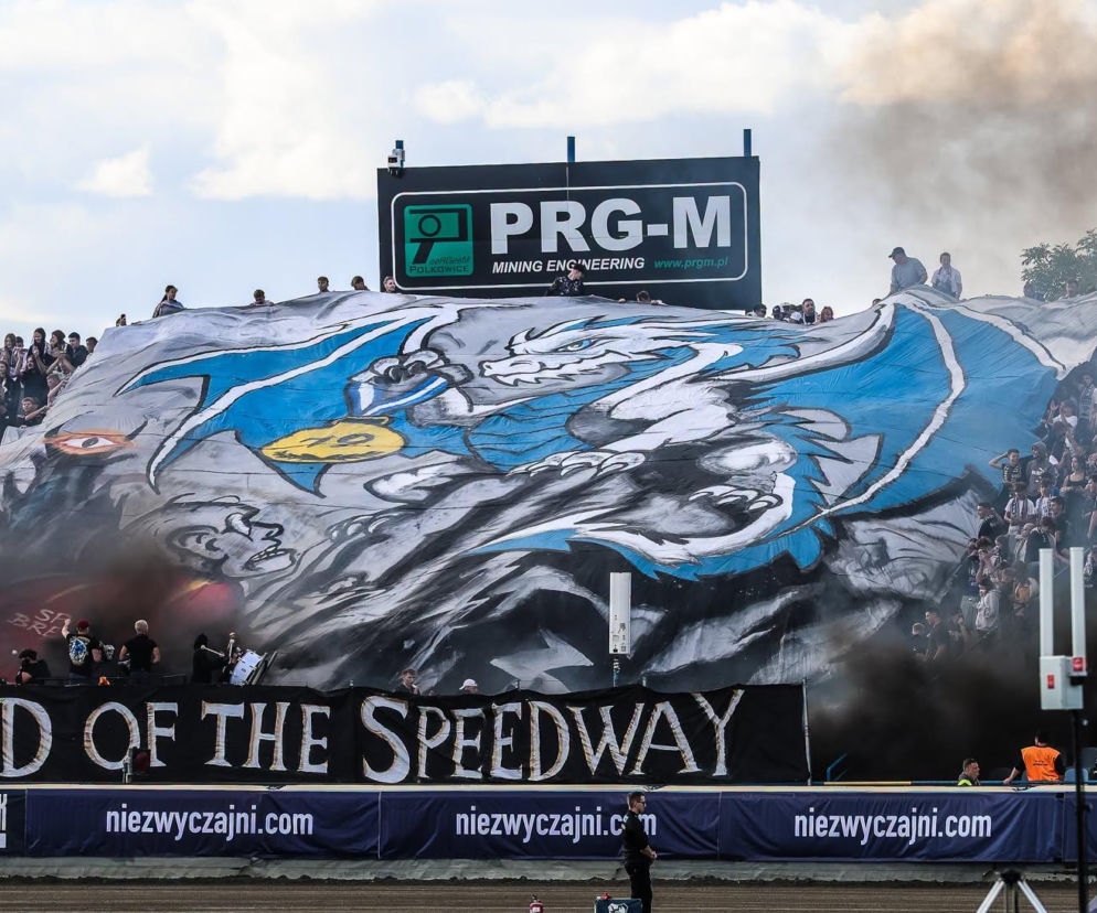 Kibice na stadionie z wielką flagą przedstawiającą smoka i napisem LORD OF THE SPEEDWAY. O Unii Leszno czytaj na Eska Leszno.