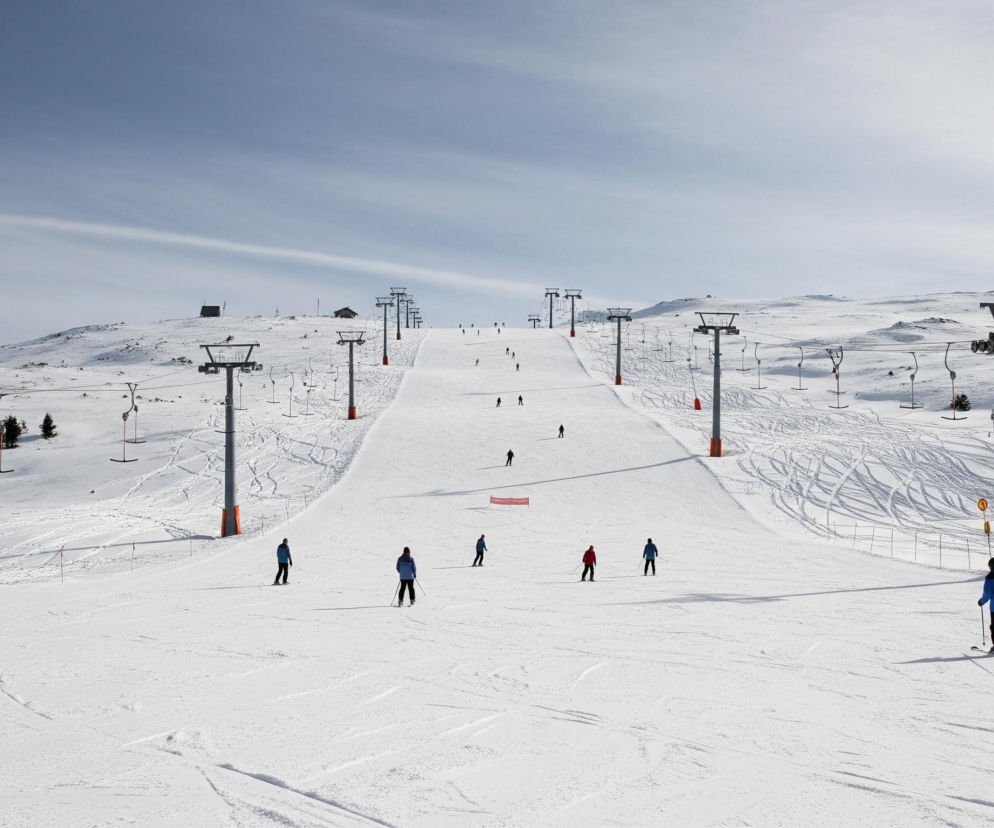 Ośnieżony stok narciarski rozciąga się pod częściowo zachmurzonym, jasnoniebieskim niebem, które zajmuje górną połowę obrazu. Wzdłuż stoku biegnie wyraźnie zaznaczone, szerokie pasmo przygotowanego śniegu, a po jego obu stronach widać liczne koleje linowe z metalowymi słupami i podwieszonymi krzesełkami lub orczykami. Na stoku widocznych jest kilkunastu narciarzy ubranych w kurtki w odcieniach niebieskiego, czerwonego i czarnego, zjeżdżających w dół lub poruszających się wzdłuż wyciągu, zostawiając za sobą ślady na białym śniegu. Na tle gór, w oddali, widać kilka niewielkich budynków, a po lewej stronie stoku, pomiędzy słupami wyciągów, rozsiane są pojedyncze, małe drzewa iglaste.