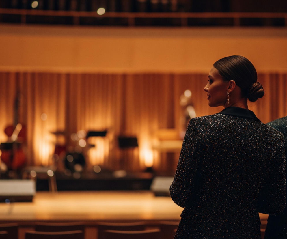 Dwie osoby, kobieta i mężczyzna, stoją tyłem do widza, obserwując scenę muzyczną. Kobieta po lewej ma ciemne włosy związane w kok, nosi długie, zwisające kolczyki oraz elegancką marynarkę w ciemnym kolorze z drobnymi, błyszczącymi elementami. Mężczyzna po prawej, widoczny od tyłu, również ma ciemną marynarkę, podobnie mieniącą się. W tle widać rozmazaną scenę z instrumentami muzycznymi, takimi jak kontrabas i perkusja, oświetloną ciepłym, pomarańczowo-złotym światłem, a za nią ścianę z pionowymi panelami w podobnym odcieniu.