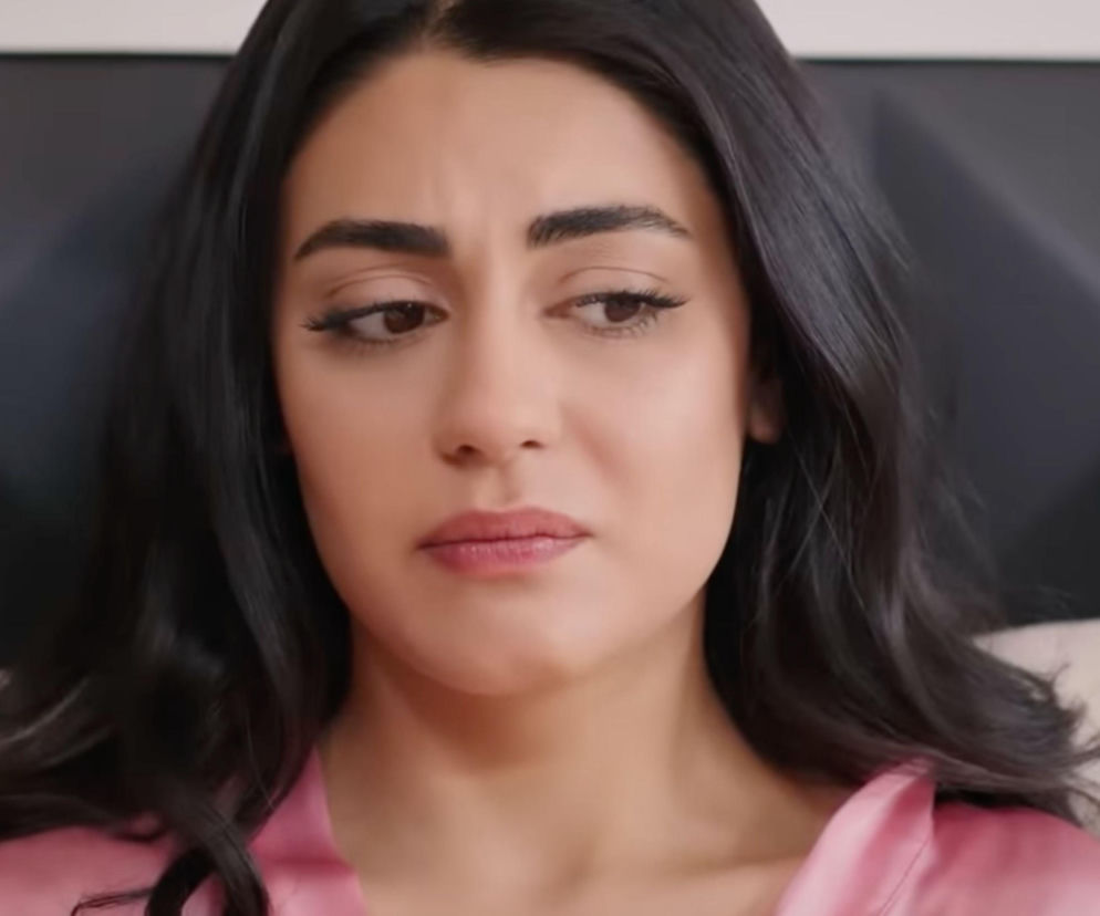 Kobieta o długich, ciemnych włosach w różowej bluzce z dekoltem w serek, z delikatnym makijażem, z powagą spogląda w dół, odzwierciedlając emocje związane z nadchodzącymi odcinkami tureckiej telenoweli „Panna młoda” na portalu Super Seriale.