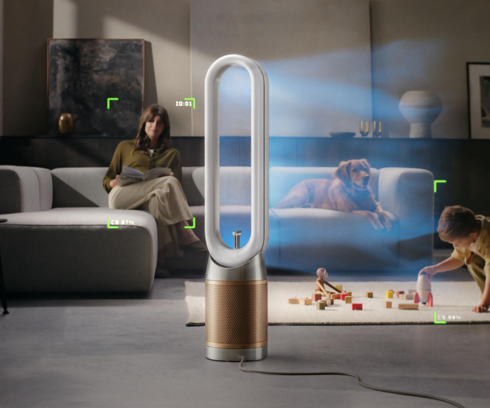 Oczyszczacz powietrza Dyson Find+Follow™ Purifier Cool w salonie, kierujący strumienie oczyszczonego powietrza do kobiety czytającej na sofie i dziecka bawiącego się na dywanie. Pies leżący na drugiej sofie również objęty jest działaniem urządzenia. Więcej o nowościach Dyson przeczytasz na naszym portalu.