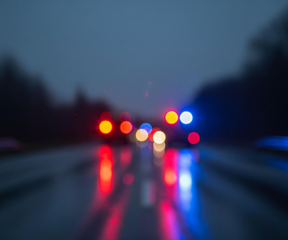 Rozmyty widok drogi w nocy lub o zmierzchu, z dominującymi odcieniami szarości i granatu w tle. Na pierwszym planie widoczne są światła pojazdów, rozmazane w okrągłe plamy, tworzące efekt bokeh. Dominują czerwone, pomarańczowe i niebieskie światła, odbijające się na mokrej nawierzchni jezdni, co tworzy wydłużone smugi kolorów. Po obu stronach drogi zarysowują się ciemne, rozmyte sylwetki drzew lub innych obiektów, zlewające się z niebem.