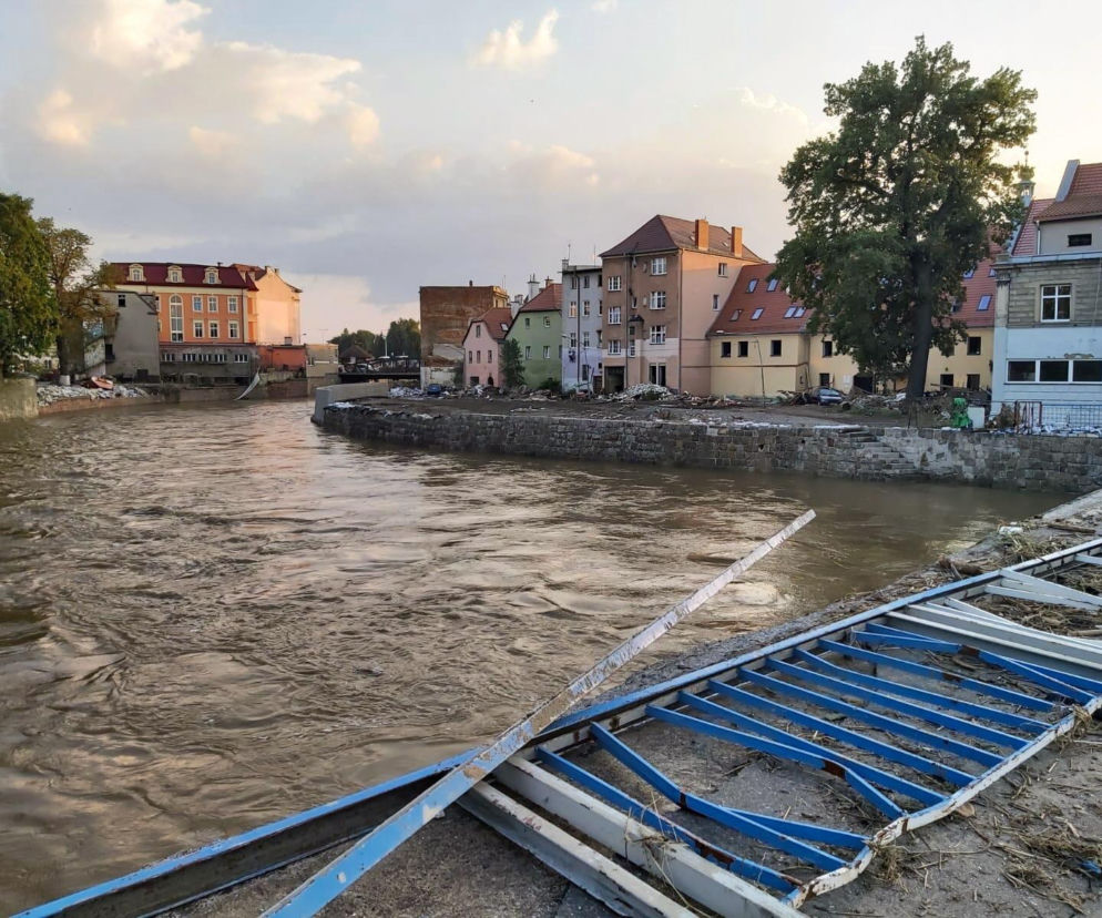 Zalana rzeka w Kłodzku po powodzi, z brązową, wzburzoną wodą i zniszczonymi budynkami na brzegu. Elementy niebieskiego ogrodzenia leżą na pierwszym planie. Na Poradnik Zdrowie znajdziesz komunikaty na temat wody.