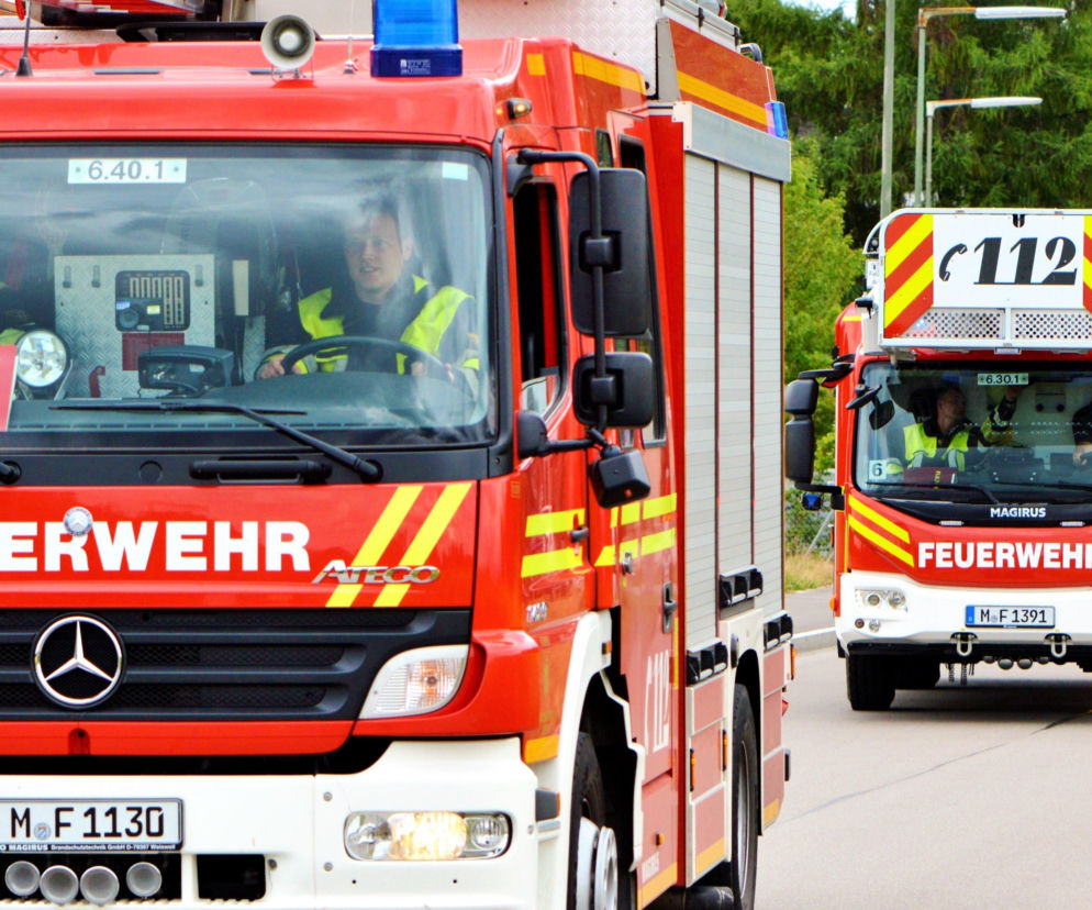 Czerwone wozy strażackie z napisem FEUERWEHR i niebieskimi światłami sygnalizacyjnymi na górze, z widocznymi strażakami w kabinach. Scena ta odzwierciedla akcję służb po pożarze ciężarówki w Niemczech, o czym więcej przeczytasz na naszym portalu.