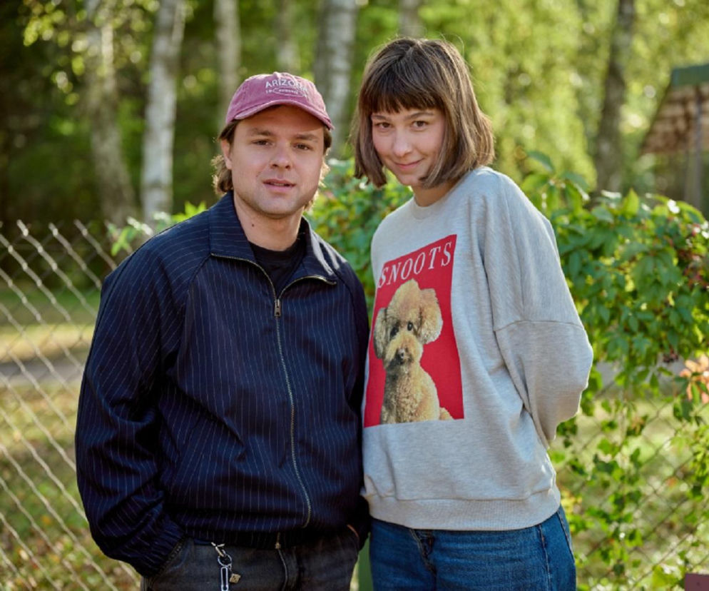 Aktorzy Maciej Kobiela jako Klusek i Maja Wolska jako Brygida z serialu „Na Wspólnej” stoją razem, patrząc wprost. Klusek w granatowej kurtce i bejsbolówce, Brygida w szarej bluzie z psem na tle zieleni. Więcej o ich losach na Super Seriale.