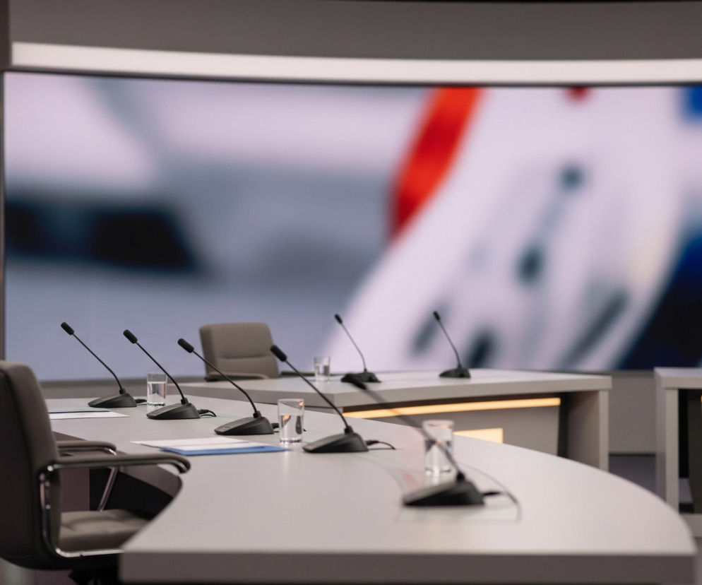 Na pierwszym planie widoczny jest szeroki, zaokrąglony stół w kolorze jasnoszarym, z rzędem pięciu czarnych mikrofonów na wysięgnikach i trzema szklankami wody. Przy stole stoją dwa ciemnoszare, obrotowe krzesła z chromowanymi podłokietnikami, skierowane tyłem do widza. W tle znajduje się duży ekran wyświetlający rozmyty obraz z elementami w kolorach białym, czerwonym i niebieskim. Po lewej stronie ekranu widać ciemne panele ścienne z poziomymi pasami jasnego podświetlenia. Po prawej stronie, za głównym stołem, znajduje się mniejszy, prostokątny stół z dwoma krzesłami, również w kolorach szarym i czarnym, oraz z mikrofonem i szklanką. Całość sprawia wrażenie nowoczesnego studia telewizyjnego lub sali konferencyjnej.