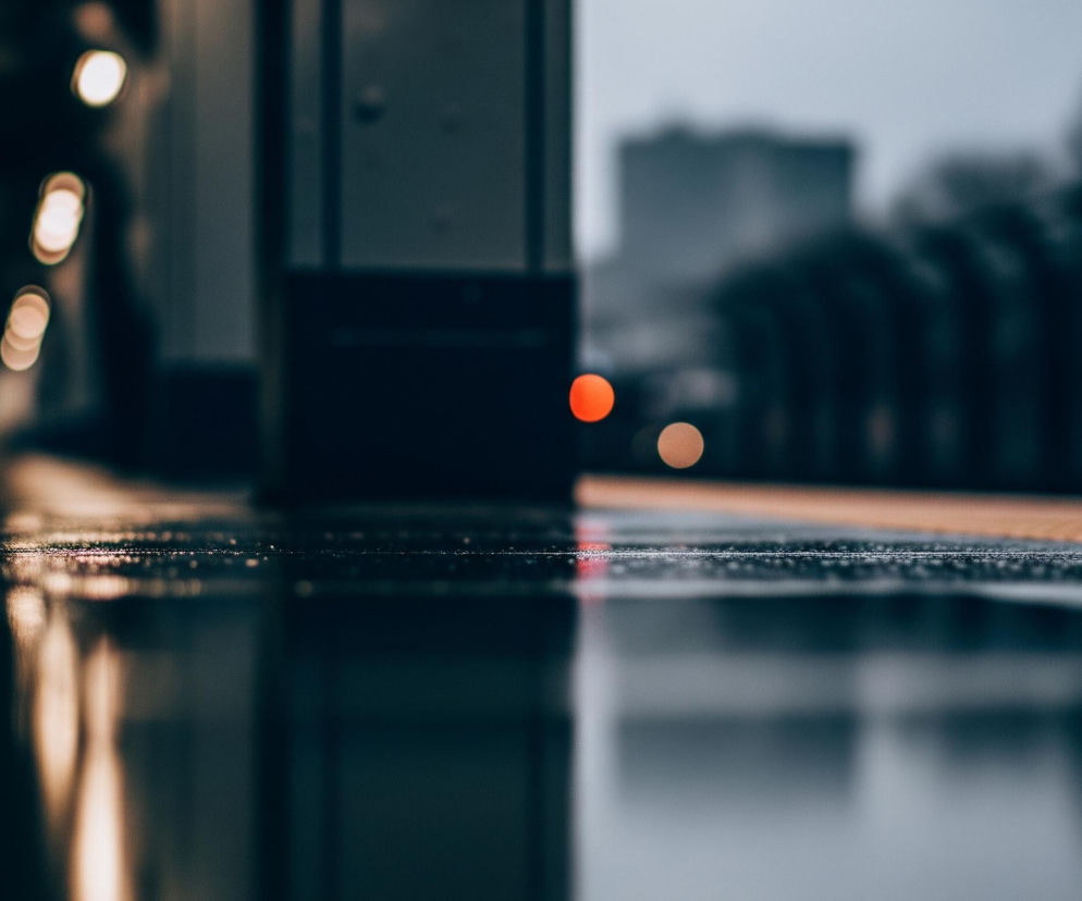 Wilgotna, ciemna powierzchnia ziemi, prawdopodobnie peron, rozciąga się od pierwszego planu do horyzontu. Po lewej stronie widać rozmyte, pionowe, jasne światła odbijające się w mokrej nawierzchni, a także pojedyncze, okrągłe, pomarańczowe światło. Po prawej stronie, w oddali, znajduje się ciemna, pozioma linia o żółtym lub pomarańczowym kolorze, a nad nią rozmyte kształty drzew i budynków pod szarym niebem. Obraz utrzymany jest w zimnych odcieniach, z wyjątkiem ciepłych punktów świetlnych.