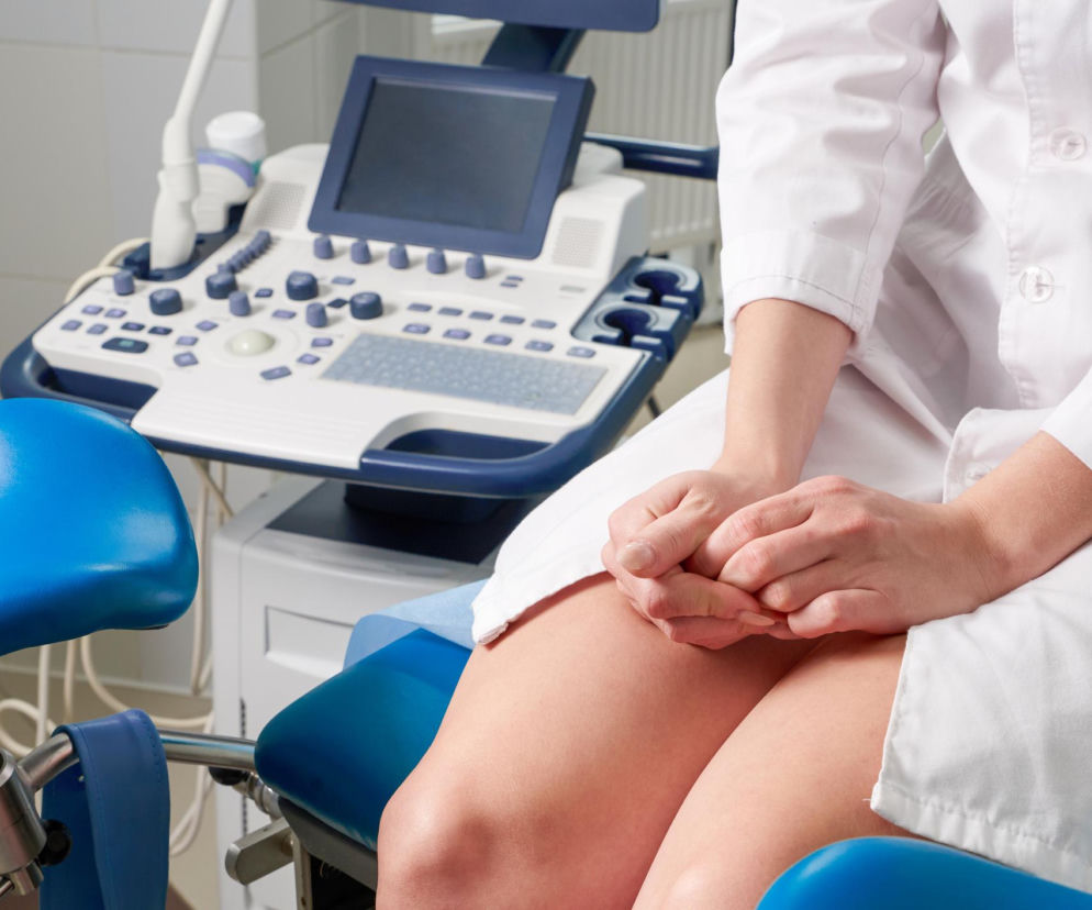 Kobieta w białym fartuchu siedząca na fotelu ginekologicznym ze splecionymi rękami, co symbolizuje niepokój przed wizytą. W tle widoczny aparat do USG. Na Poradnik Zdrowie znajdziesz informacje o profilaktyce nowotworów ginekologicznych.