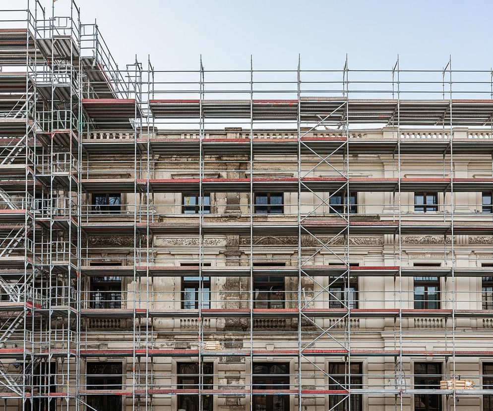 Monumentalna fasada budynku jest niemal w całości zasłonięta przez złożone rusztowanie konstrukcyjne wykonane z metalowych rur i drewnianych podestów. Centralna część budynku, widoczna między elementami rusztowania, ukazuje elewację w jasnym, beżowym kolorze, zdobioną bogatymi, ciemnymi ornamentami i prostokątnymi oknami z ciemnymi ramami. Niebo ponad budynkiem jest jasnoniebieskie, bezchmurne.