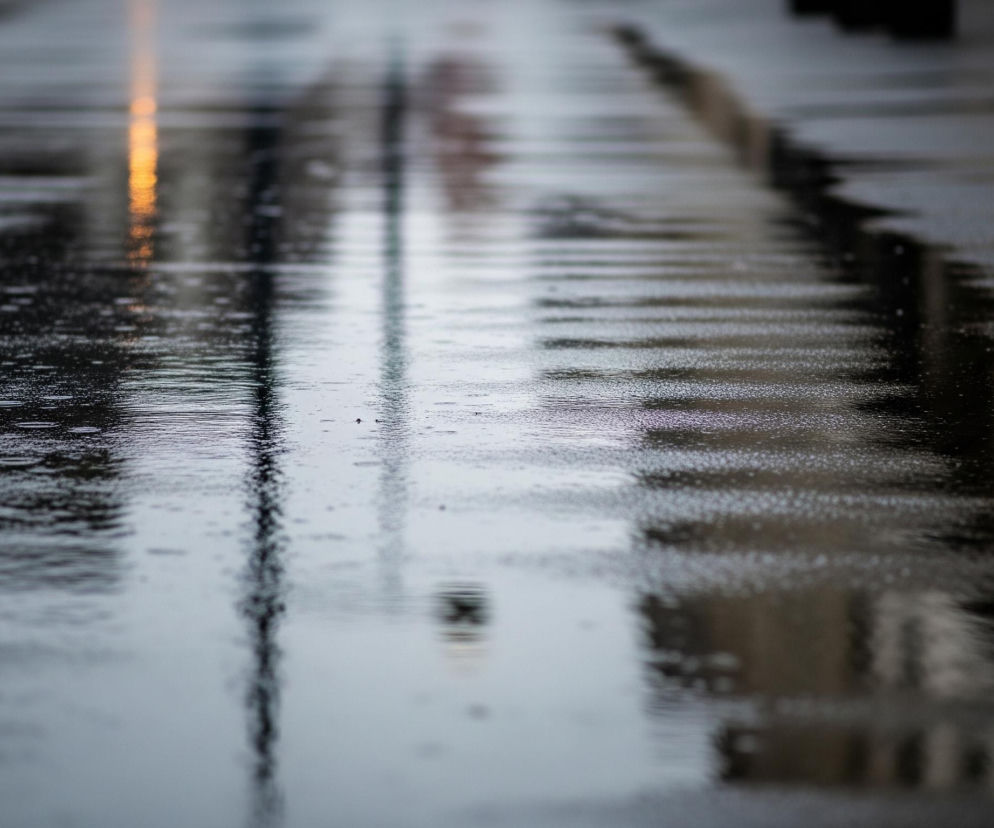 Mokra, ciemnoszara nawierzchnia drogi lub chodnika odbija światło, tworząc refleksy. Po lewej stronie w górnej części widać wydłużony, pionowy, rozmyty, jasnopomarańczowy refleks, prawdopodobnie lampy ulicznej. W centralnej części obrazu dominują pionowe, rozmyte cienie słupów lub drzew, a po prawej stronie zarys ciemnych budynków. Na całej powierzchni widoczne są drobne kropelki deszczu i zmarszczki na tafli wody, świadczące o trwającym opadzie.