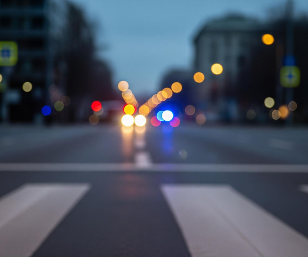 Przedstawia rozmyty widok ulicy miejskiej o zmierzchu lub w nocy, z ostrością ustawioną na pierwszoplanowe pasy dla pieszych. Na pierwszym planie, w dolnej części kadru, widoczne są białe pasy przejścia dla pieszych na ciemnoszarym asfalcie, które biegną ukośnie od lewej do prawej strony. W oddali, w centralnej części zdjęcia, skupione są jasne, rozmyte światła samochodów i latarni ulicznych w odcieniach pomarańczowego, czerwonego, niebieskiego i białego, tworząc efekt bokeh. Tło stanowią rozmazane, ciemne sylwetki budynków i drzew pod pochmurnym, ciemnoniebieskim niebem.