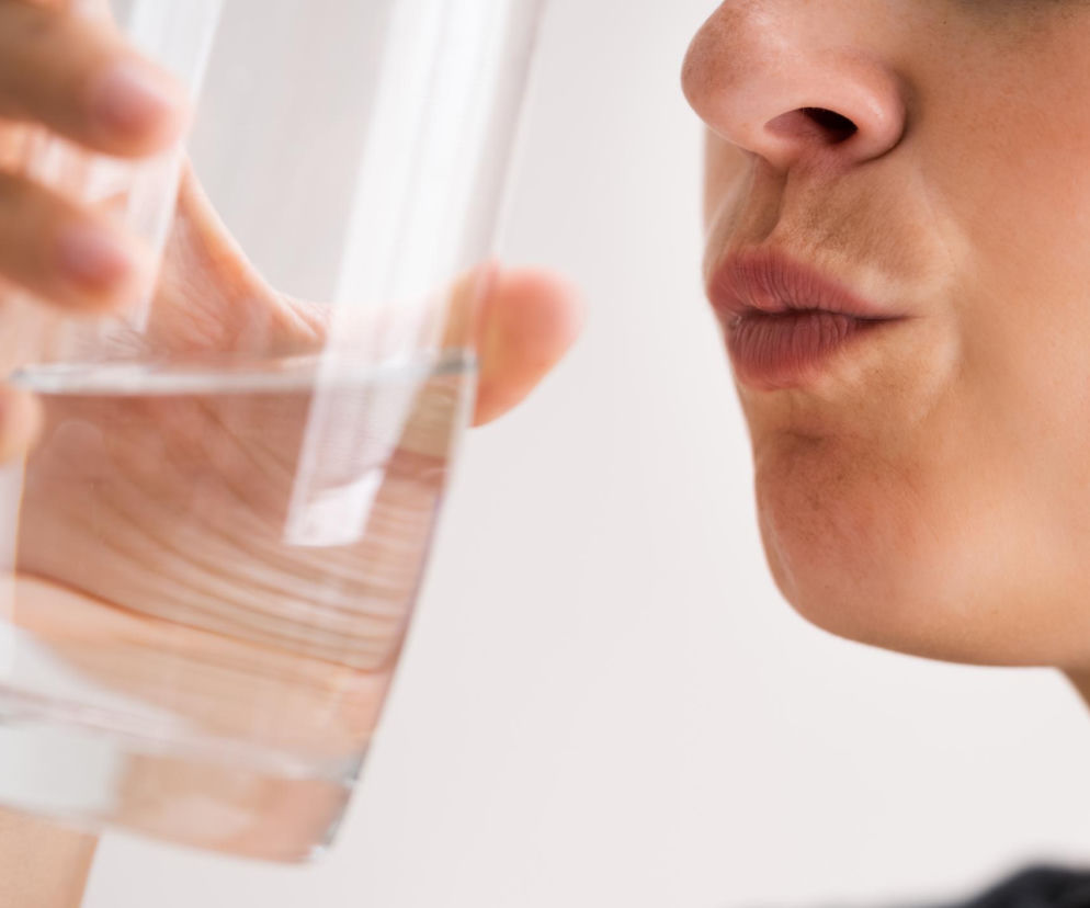 Kobieta pije wodę ze szklanki, wyraźnie widoczne usta i nos. Scena symbolizuje odświeżenie lub próbę pozbycia się nieprzyjemnego smaku. Artykuł na Poradnik Zdrowie wyjaśnia, co oznacza metaliczny posmak w ustach.