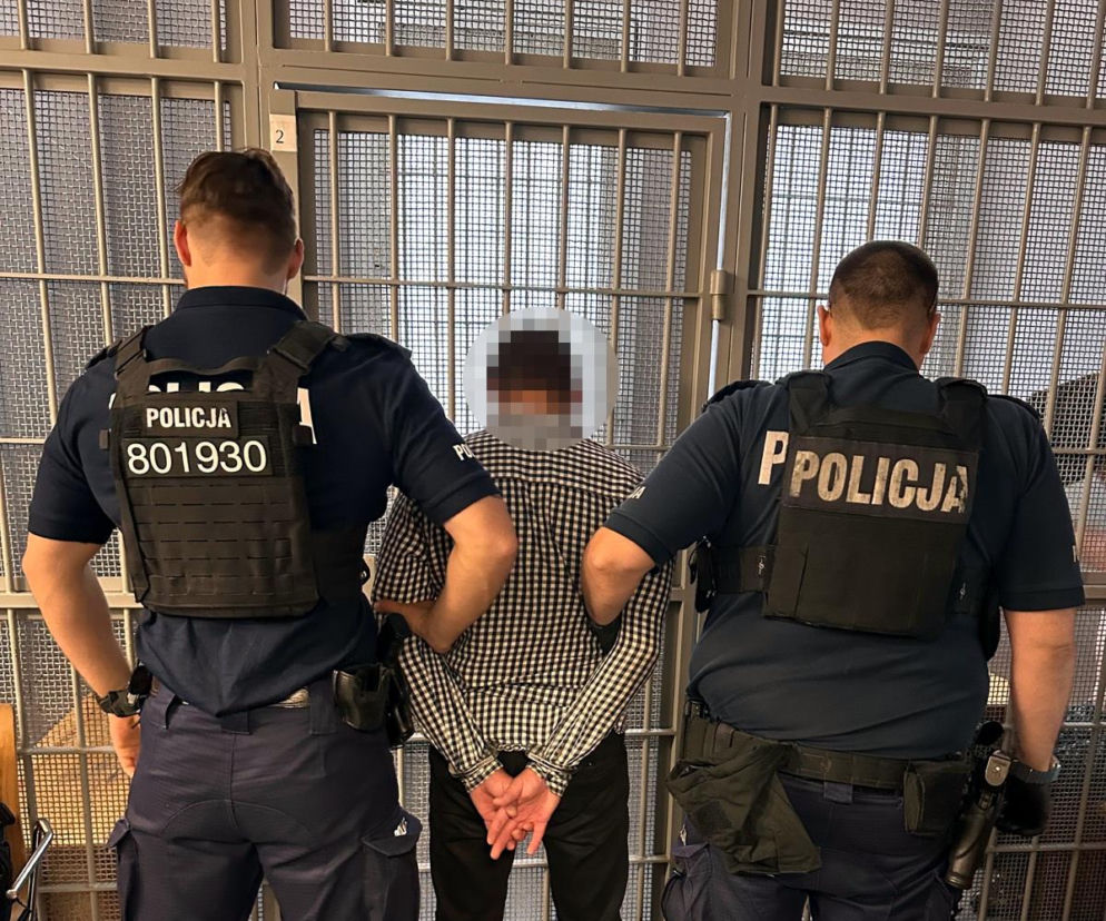 Dwaj policjanci trzymają za ręce zatrzymanego mężczyznę w kratę przed kratami aresztu. O sprawie przeczytasz na SE.
