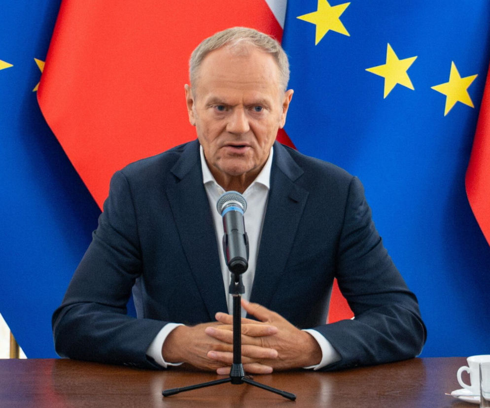 Premier Donald Tusk przy stole z mikrofonem na tle flag Polski i Unii Europejskiej. O zmianach w rządzie pisze SE Polityka.