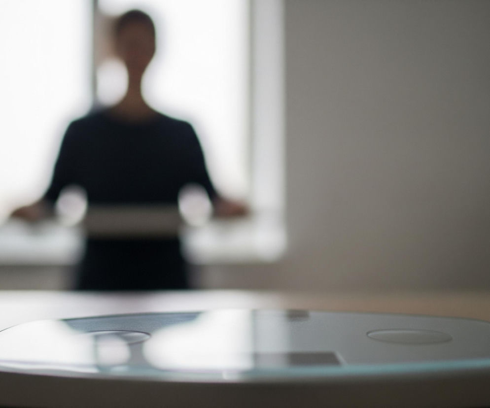 Na pierwszym planie, w centrum, znajduje się biała, płaska waga łazienkowa z zaokrąglonymi krawędziami, której górna powierzchnia jest wykonana z odblaskowego materiału, być może szkła, odbijającego światło z góry. Waga leży na jasnobrązowym, drewnianym lub laminowanym blacie. W tle, za blatem, widoczna jest niewyraźna sylwetka osoby stojącej przed dużym, jasnym oknem, którego światło tworzy poświatę. Osoba ma na sobie ciemne ubranie i trzyma coś w rękach na wysokości talii.