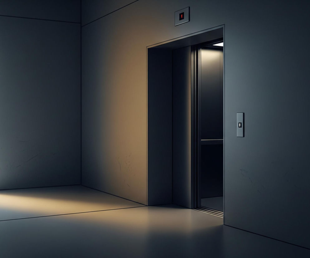 Ciemny korytarz z drzwiami windy po prawej stronie. Drzwi windy są lekko otwarte, ukazując ciemne wnętrze, a nad nimi widać prostokątny panel z czerwonym, świecącym kwadratem. Po prawej stronie drzwi windy, na ścianie, umieszczony jest pionowy panel z przyciskiem. Lewa część korytarza jest jaśniejsza, z narożnikiem ściany i fragmentem podłogi oświetlonym jasnym, białym światłem, które przechodzi w ciepłe, złociste odcienie w kierunku drzwi windy. Cała przestrzeń utrzymana jest w chłodnej palecie barw, dominują szarości i ciemne granaty, przełamane jasnym oświetleniem po lewej stronie.