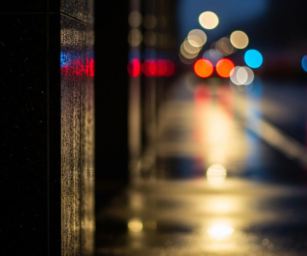 Lewa strona kadru ukazuje pionową, ciemną, chropowatą powierzchnię, pokrytą drobnymi kroplami wody, które odbijają rozproszone światła w kolorach czerwonym i niebieskim. Powierzchnia ta tworzy ciemne tło w tle, podczas gdy prawa strona zdjęcia przedstawia rozmazane, okrągłe światła bokeh w odcieniach czerwieni, bieli, błękitu i ciepłego żółtego, sugerujące miejski krajobraz nocą. Te światła rzucają podłużne refleksy na mokrej, ciemnej nawierzchni.