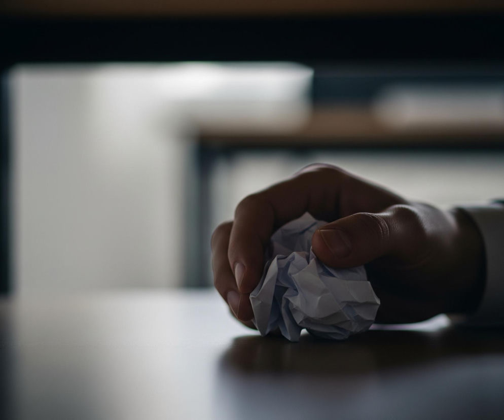 Prawa ręka osoby dorosłej ściska pomiętą kartkę białego papieru, leżącą na ciemnym, błyszczącym blacie stołu. Osoba ma na sobie ciemny garnitur i białą koszulę z długim rękawem. W tle widać niewyraźne zarysy mebli, prawdopodobnie nóg stołu i innych powierzchni. Oświetlenie jest słabe, tworząc cienie i odbicia na blacie.