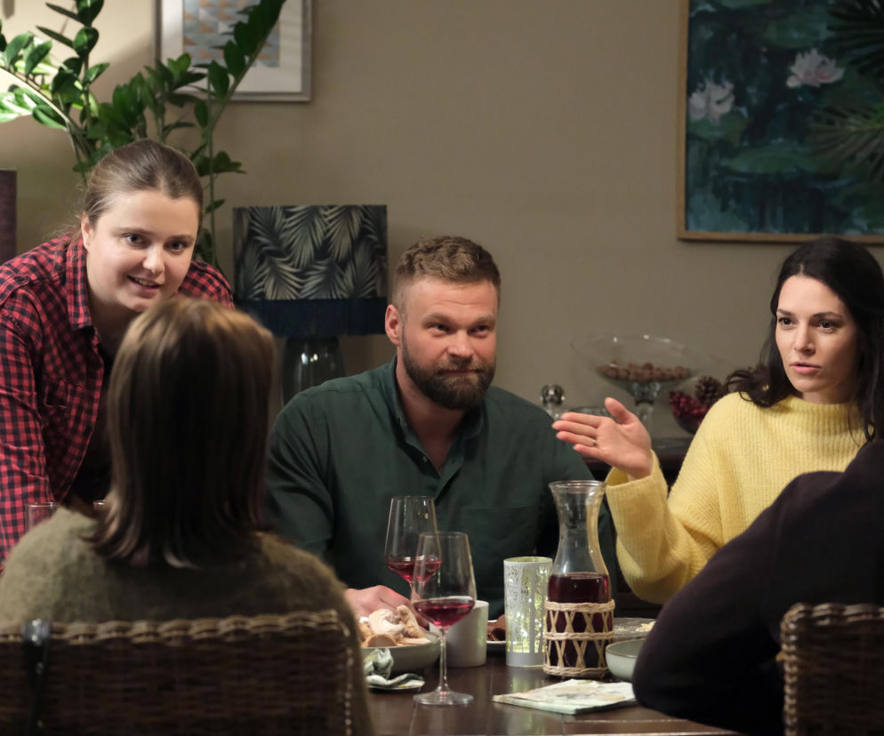 Ewelina (Anna Jarosik) i Michał (Tomasz Rożniatowski) przy stole podczas parapetówki. Ewelina w żółtym swetrze gestykuluje, rozmawiając z gośćmi, a Michał w zielonej koszuli z brodą patrzy w lewo. Na stole jedzenie, wino i lampka, atmosfera spotkania przyjaciół. Więcej o ich losach na Super Seriale.