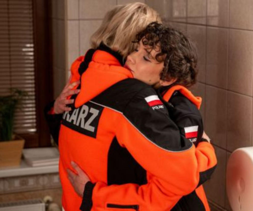 Dwie kobiety w pomarańczowo-czarnych kurtkach ratowników medycznych obejmują się, jedna z nich ma krótkie, kręcone ciemne włosy i opiera głowę na ramieniu drugiej, blondynki. Na kurtkach widać napisy KARZ i flagę Polski. Więcej o ich losach w Na sygnale przeczytasz na portalu Super Seriale.