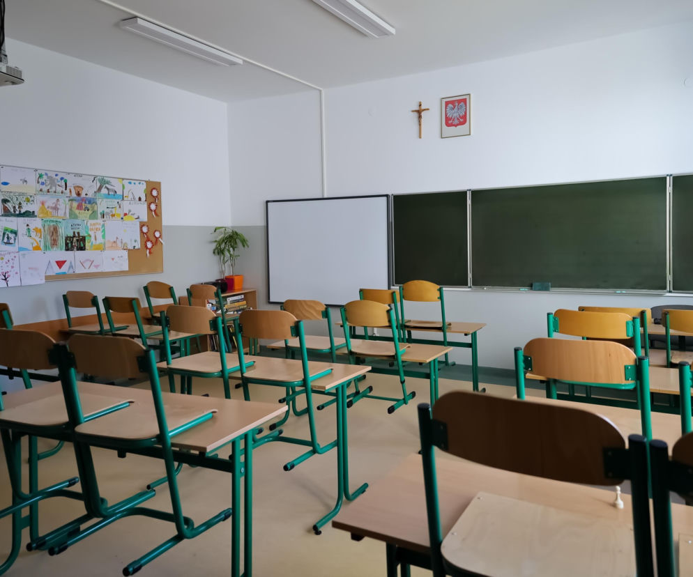 Szkolna klasa z ławkami i tablicą, nad którą wisi krzyż i godło. O decyzji MEN w SE Lublin.