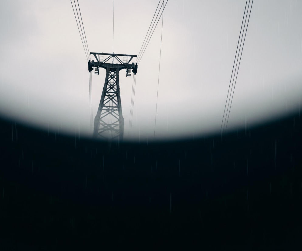 Na pierwszym planie, w dolnej części obrazu, widoczna jest ciemna, niemal czarna masa, która lekko zakrzywia się ku górze, tworząc miękki łuk. Na tej ciemnej powierzchni oraz w górnej części obrazu, widoczne są drobne, pionowe smugi, imitujące padający deszcz.

W tle, dominującym elementem jest ciemna, metalowa konstrukcja przypominająca słup kolejki linowej, wyróżniająca się na tle jasnego, zamglonego nieba. Słup ma kratownicową konstrukcję, a na jego szczycie znajdują się poziome elementy z kołami, od których odchodzą równoległe, cienkie liny, przecinające górną część kadru. Całość utrzymana jest w stonowanej kolorystyce szarości, czerni i bieli.