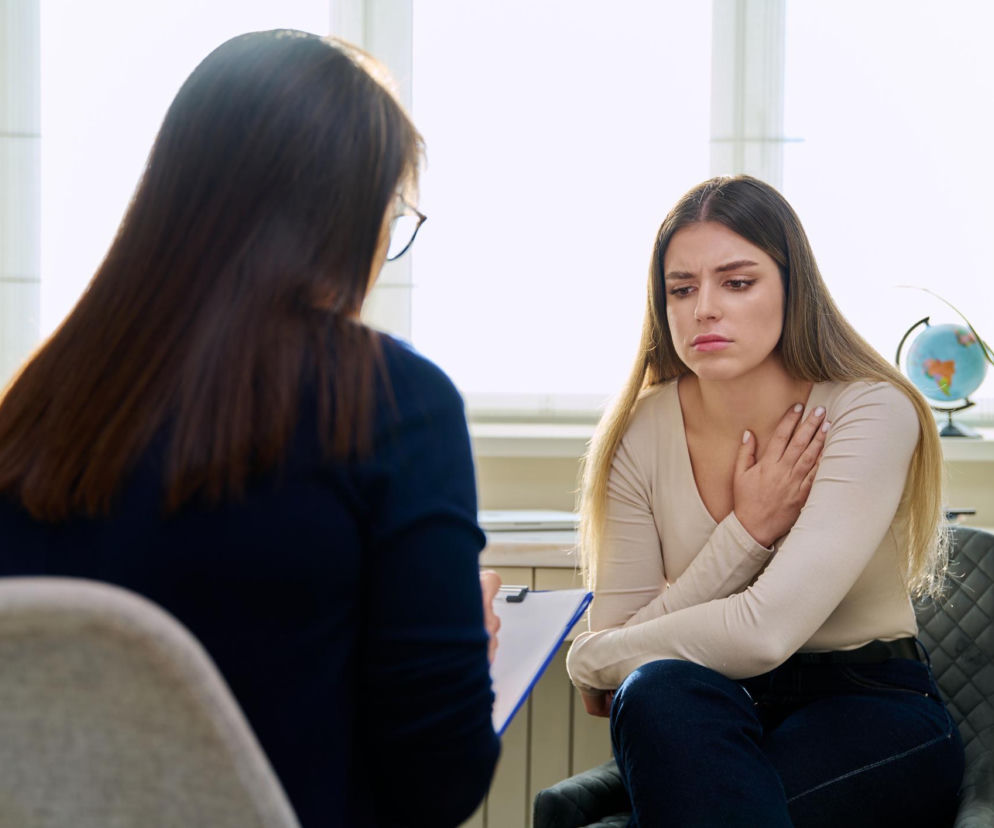 Kobieta podczas sesji z psychologiem, siedząca na krześle, z dłonią na klatce piersiowej, wyraźnie zaniepokojona. Specjalista siedzi tyłem, z notatnikiem w ręku. Artykuły na Poradnik Zdrowie obalają mity dotyczące psychologów i wskazują drogę do zdrowia psychicznego.