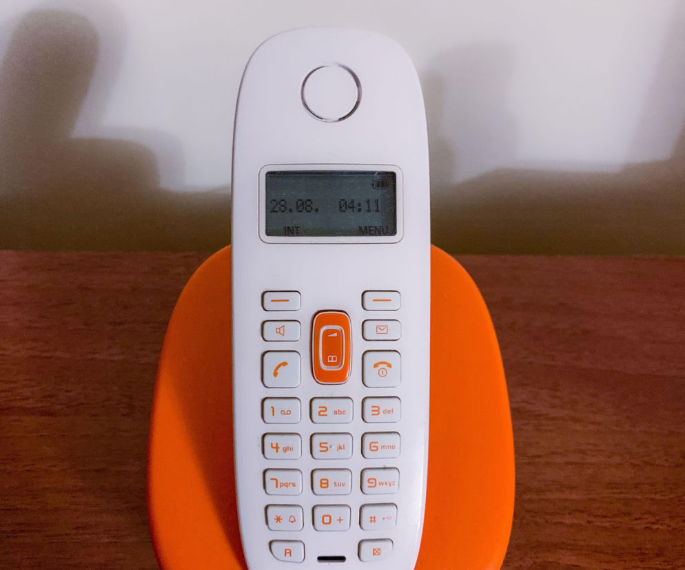 Biały, bezprzewodowy telefon stacjonarny z pomarańczowymi przyciskami i wyświetlaczem z datą i godziną, umieszczony na pomarańczowej podstawce ładującej na drewnianym blacie. Obraz symbolizuje koniec ery telefonii stacjonarnej w Finlandii, o czym przeczytasz więcej na naszym portalu.