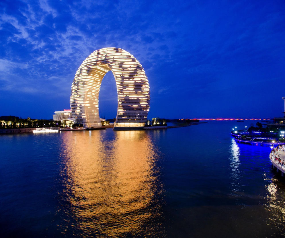Hotel Sheraton Huzhou Hot Spring Resort w Huzhou w Chinach w kształcie pierścienia, oświetlony w nocy, z odbiciami świateł na tafli jeziora. To architektoniczne arcydzieło można podziwiać na portalu Architektura Murator Plus.