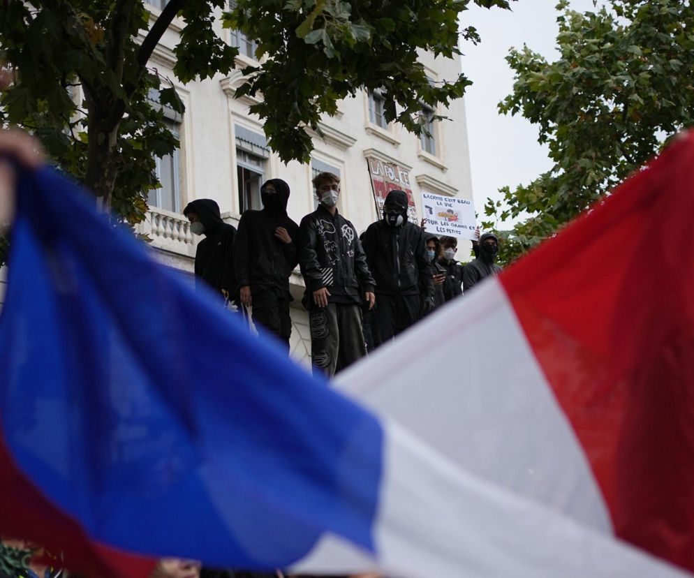 Grupa zamaskowanych osób na podwyższeniu za flagami Francji podczas protestów. O sytuacji we Francji przeczytasz na SE.