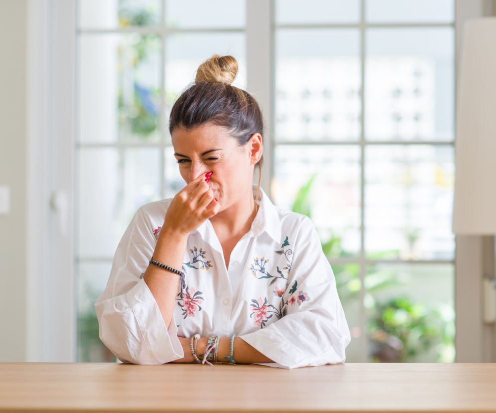 Młoda kobieta z burzą loków i upiętym kokiem, w białej koszuli z kwiatowymi haftami, siedzi przy drewnianym stole i zasłania nos palcami, marszcząc twarz z niezadowolenia. Ilustracja symbolizuje nieprzyjemny zapach potu, o którego przyczynach przeczytasz na Poradnik Zdrowie.