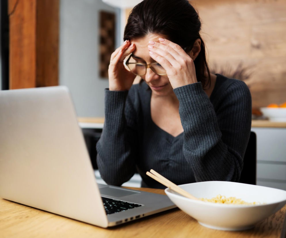 Kobieta w okularach, z dłońmi na skroniach, siedzi przy stole z laptopem i miską makaronu z pałeczkami, symbolizując mgłę mózgową po jedzeniu. Artykuł na Poradniku Zdrowie wyjaśnia, jak dieta i stan jelit wpływają na koncentrację i zmęczenie.