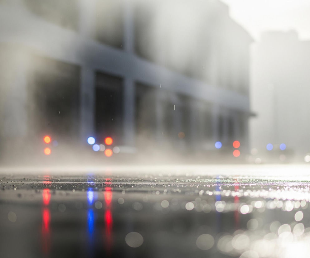 Szeroki kadr przedstawia perspektywę niskiego kąta na mokrą powierzchnię ulicy lub placu, z wyraźnymi kroplami wody i odbiciami światła tworzącymi bokeh w dolnej części obrazu. W tle, rozmyte budynki o szaro-beżowych odcieniach, z widocznymi, niewyraźnymi oknami i fasadami, przechodzą w jasne, niemal białe niebo. Pośrodku obrazu, w tle, pojawiają się rozmyte światła w kolorach czerwonym i niebieskim, które odbijają się także na mokrej nawierzchni w dolnej części kadru. Ogólna atmosfera jest zamglona i wilgotna, z widocznymi pojedynczymi liniami deszczu lub pary unoszącej się w powietrzu.