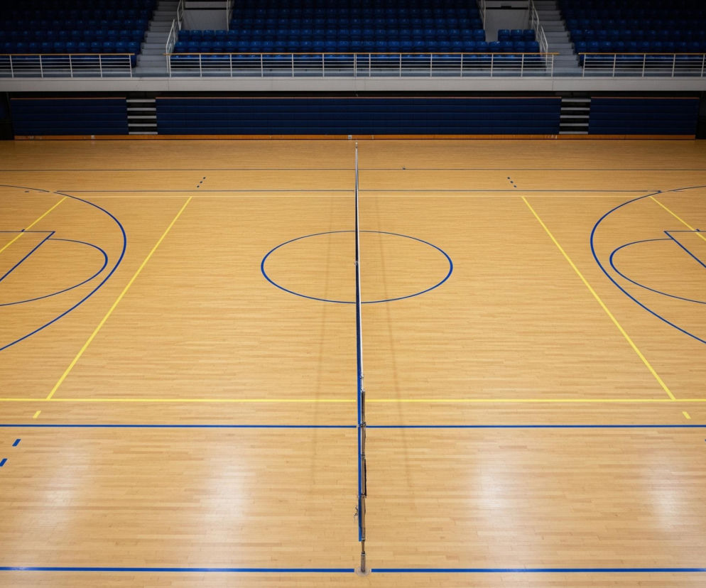 Na pierwszym planie widoczny jest jasny, drewniany parkiet boiska sportowego z wyraźnymi niebieskimi i żółtymi liniami. Centralnie umieszczona jest siatka do siatkówki, dzieląca boisko na dwie równe części, z okrągłym, niebieskim okręgiem dokładnie na środku. W tle, powyżej boiska, rozciągają się rzędy ciemnoniebieskich siedzeń na trybunach, częściowo oświetlone, a częściowo pogrążone w cieniu, otoczone białymi barierkami.