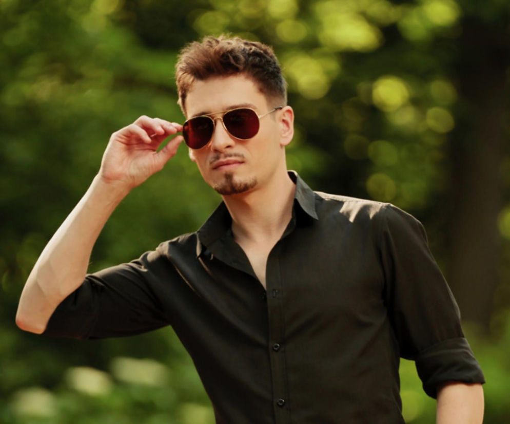 Mateusz Śniechowski (MATEO) w czarnej koszuli i okularach przeciwsłonecznych, pozujący na tle zielonej roślinności. Zdjęcie ilustruje artykuł o jego nowym klipie i karierze muzycznej, o czym więcej przeczytasz na naszym portalu.
