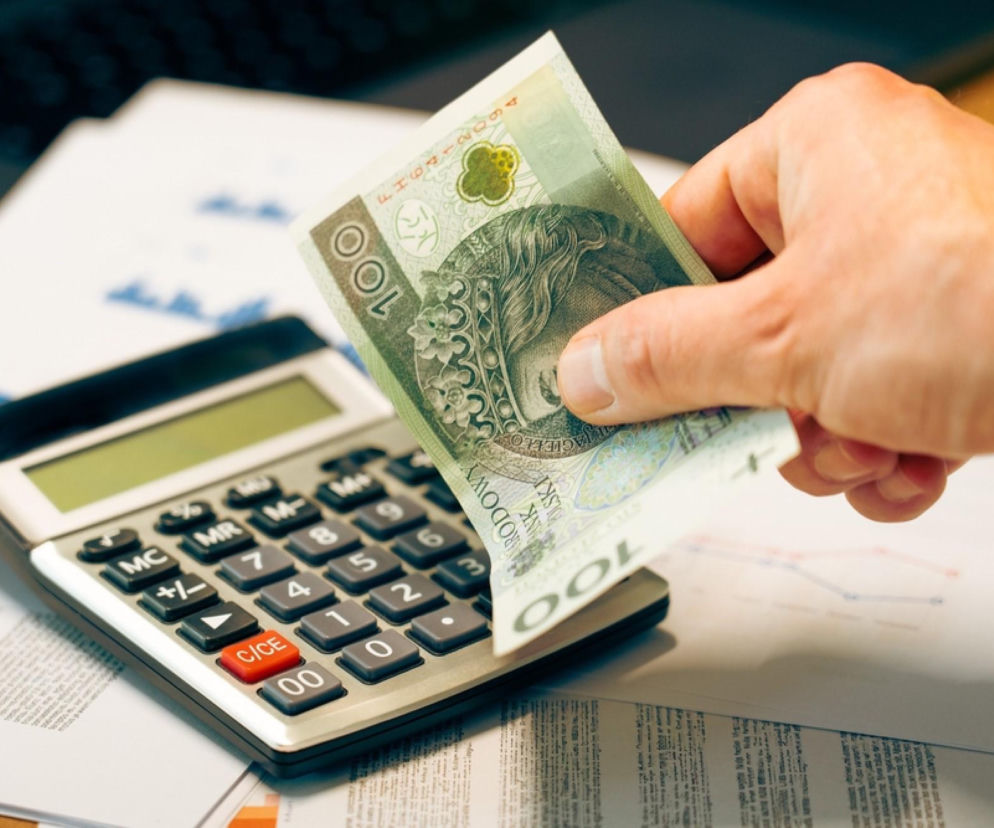 Dłoń trzymająca banknot 100 zł nad kalkulatorem i dokumentami finansowymi, symbolizująca rozliczenia podatkowe. Zdjęcie ilustruje temat płatności podatku od nieruchomości do 15 marca, o którym można przeczytać na Super Biznes.