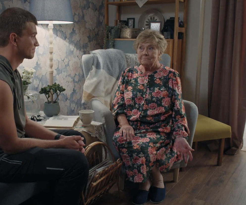 Rafał Kowalski (Mateusz) siedzi na wprost Teresy Lipowskiej (Barbara Mostowiak) w jej salonie, patrząc w bok z lekkim uśmiechem. Barbara w kwiecistej sukience siedzi na fotelu, spoglądając na wnuka. Oboje są w swobodnej pozycji, a obok nich znajduje się stolik z książką i filiżanką. Więcej o ich losach w M jak miłość przeczytasz na Super Seriale.