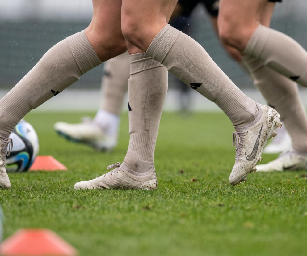 Nogi piłkarzy Rakowa Częstochowa w beżowych getrach i białych korkach z czarnym logo Nike, stojące na zielonej murawie, obok leżą dwie piłki nożne. Treningi przed sezonem Ekstraklasy są intensywne, o czym przeczytasz więcej na naszym portalu.