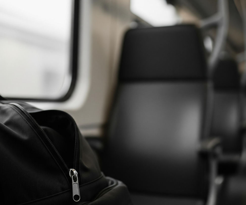Czarny plecak, częściowo rozpięty, leży na siedzeniu w pociągu, zajmując lewą część kadru. Za plecakiem widać okno z niewyraźnym, jasnym tłem, a po prawej stronie kadru, również rozmazane, znajdują się rzędy pustych, ciemnych siedzeń. Oświetlenie wewnątrz pociągu jest przygaszone, tworząc stonowaną paletę barw skupioną wokół czerni, szarości i bieli.