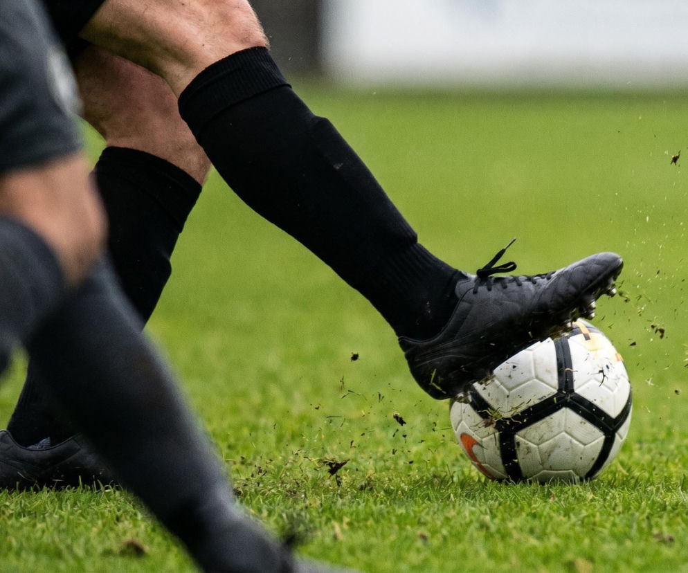 Noga piłkarza w czarnym stroju i butach uderza w piłkę na zielonej murawie, rozrzucając wokół niej trawę. Scena sportowa symbolizuje rywalizację i dynamikę, o której przeczytasz więcej na naszym portalu w kontekście faworytów MŚ 2026.