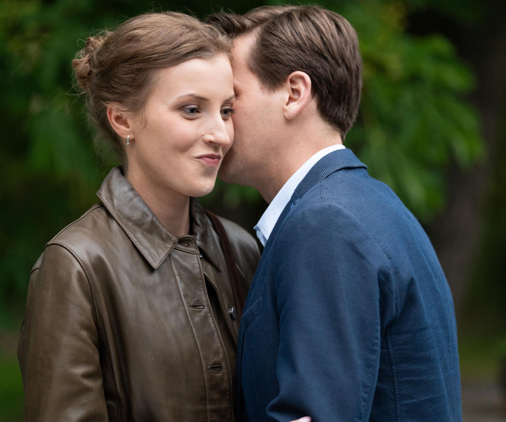 Zbliżenie Natalki (Dominika Suchecka) i Adama (Patryk Szwichtenberg) z serialu M jak miłość. Mężczyzna szepcze do ucha uśmiechniętej kobiecie, która ma na sobie brązową kurtkę. Para zbliża się do siebie, co jest tematem intryg Hani. Więcej o ich losach na Super Seriale.