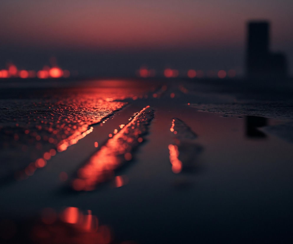 Rozlana woda z wyraźnymi smugami na ciemnej, chropowatej powierzchni zajmuje pierwszy plan, odbijając intensywne, pomarańczowo-czerwone światła z oddali. Na linii horyzontu rozciąga się pas rozmytych, jasnoczerwonych świateł, a w prawym górnym rogu zarysowuje się ciemna sylwetka budynku, również rozmyta. Cała scena jest zdominowana przez ciemne barwy, takie jak granat i czerń, przełamane mocnym pomarańczowo-czerwonym blaskiem odbitym od powierzchni i światłami w tle.