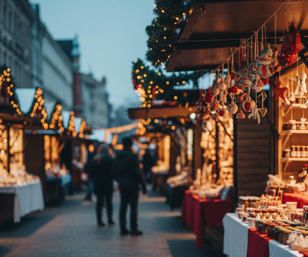 Na pierwszym planie widoczny jest stragan jarmarczny, ozdobiony girlandami ze sztucznych choinek i licznymi, ciepło świecącymi lampkami. Spod dachu straganu zwisają różnorodne, ręcznie robione ozdoby, w tym małe figurki bałwanów, czerwone serca i białe bombki, natomiast na ladzie ustawione są słoiki z przetworami i butelki z płynami. Stragany te ciągną się w głąb ulicy, tworząc aleję oświetloną żółtym blaskiem lampek, gdzie w tle widać rozmyte sylwetki ludzi, przechadzających się między stoiskami. Całość tonie w zmierzchowym, nieco niebieskawym świetle nieba, które kontrastuje z ciepłymi barwami jarmarku.