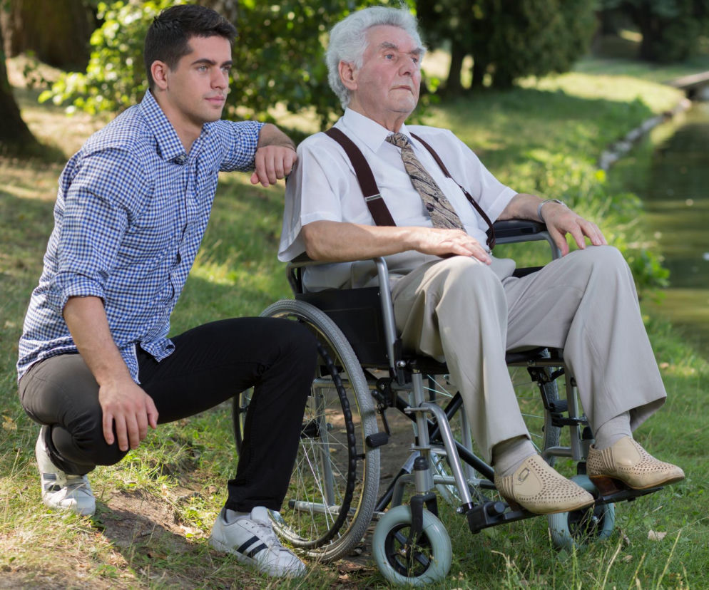 Młody mężczyzna kuca obok starszego pana na wózku inwalidzkim, patrząc w tym samym kierunku. Obaj siedzą w otoczeniu zielonego parku z wodą w tle, co może symbolizować opiekę i wsparcie w chorobie Alzheimera. Więcej o mechanizmach choroby na Poradnik Zdrowie.