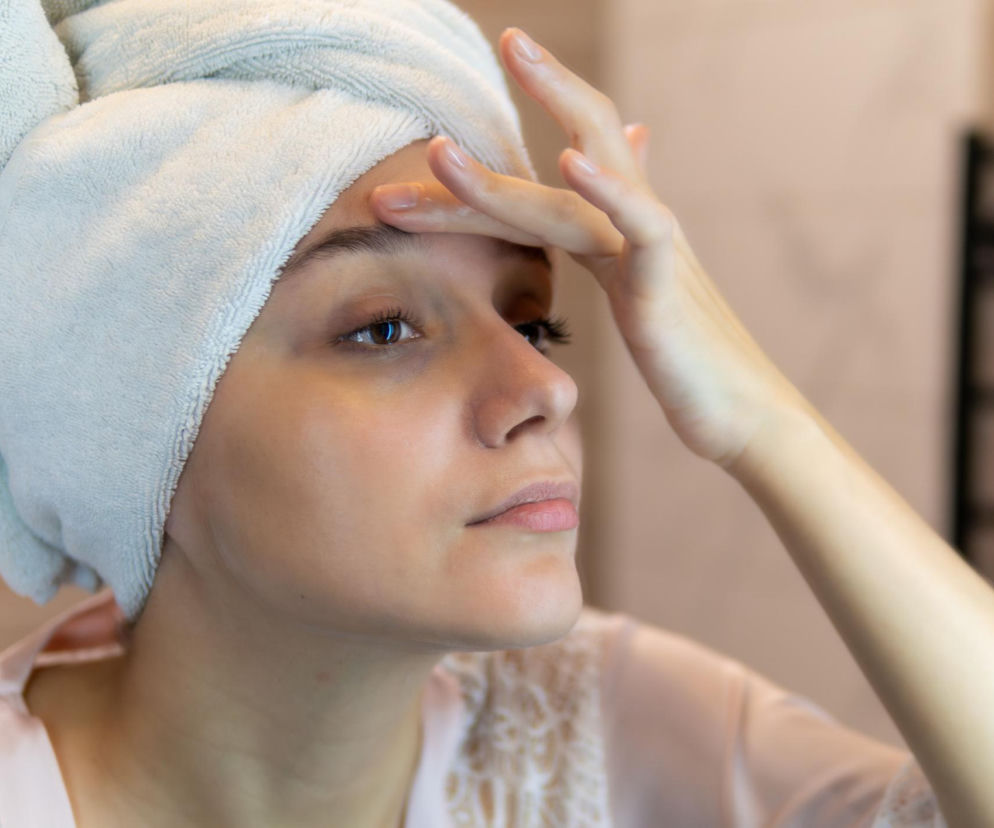 Kobieta z ręcznikiem na głowie sprawdza palcem brwi w lustrze, analizując ich stan. Objaw Hertoghe’a i wypadanie brwi mogą świadczyć o problemach z tarczycą. Więcej o chorobie Hashimoto i niedoczynności tarczycy przeczytasz na Poradnik Zdrowie.