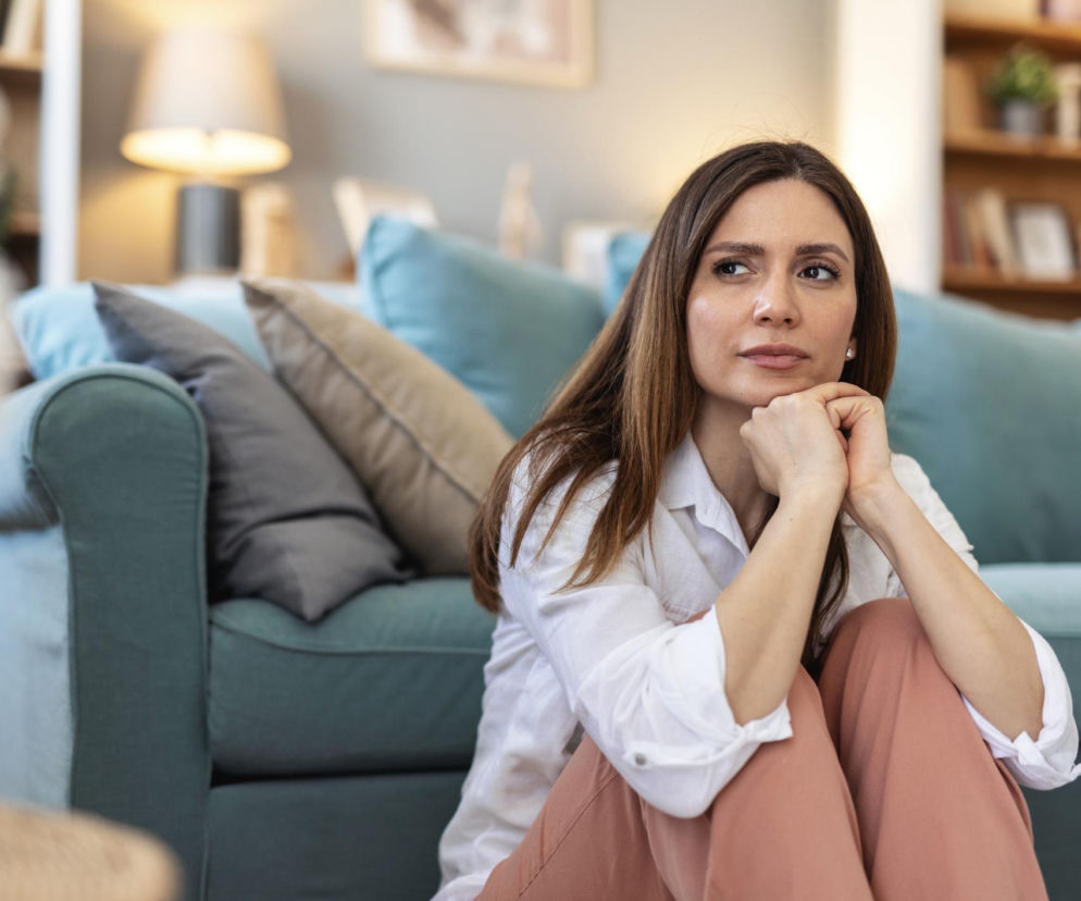 Młoda kobieta z długimi, brązowymi włosami siedząca na podłodze w salonie obok kanapy, z zamyślonym wyrazem twarzy, nawiązuje do kaca społecznego. Jej poza i wyraz twarzy sugerują zmęczenie lub introspekcję. Więcej o zjawisku zmęczenia po spotkaniach towarzyskich przeczytasz na Poradnik Zdrowie.