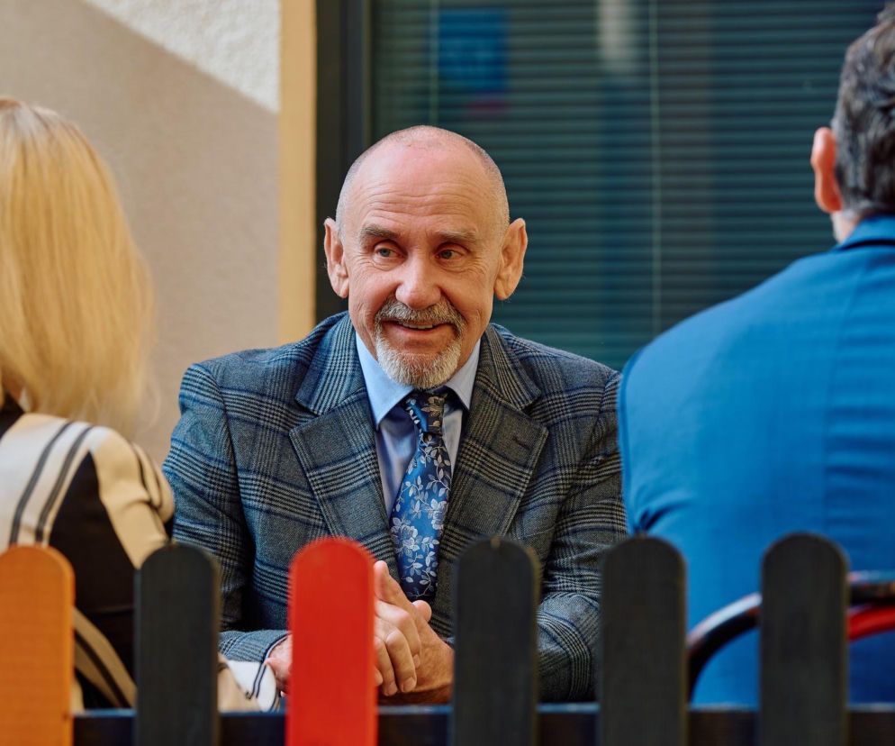 Uśmiechnięty Bożydar w kraciastej marynarce rozmawia z ludźmi przy płotku. O buncie sąsiadów przeczytasz na SE Superseriale.