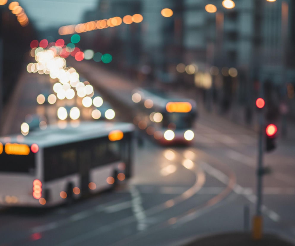 Na rozmytej fotografii miejskiej ulicy o zmierzchu, w centrum widoczne są dwa rozmyte autobusy. Pierwszy, biały z ciemnymi szybami, z tyłem skierowanym lekko w prawo, ma zapalone czerwone światła pozycyjne i pomarańczowy, podświetlony wyświetlacz na tyle. Drugi autobus, w tle po prawej stronie, jest częściowo widoczny z przodu i również ma zapalone jasne światła. Wzdłuż ulicy ciągną się rzędy rozmytych, jasnych świateł pojazdów i ulicznych lamp, tworząc bokeh w odcieniach pomarańczowego, żółtego, czerwonego i zielonego. Po prawej stronie kadru znajduje się ciemny słupek z zapalonymi czerwonymi światłami sygnalizacji świetlnej.