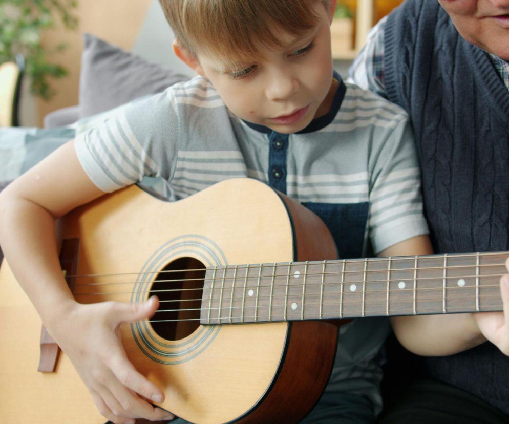 Chłopiec trzyma gitarę akustyczną na kolanach i dotyka strun. O wyborze pierwszej gitary przeczytasz na SE.