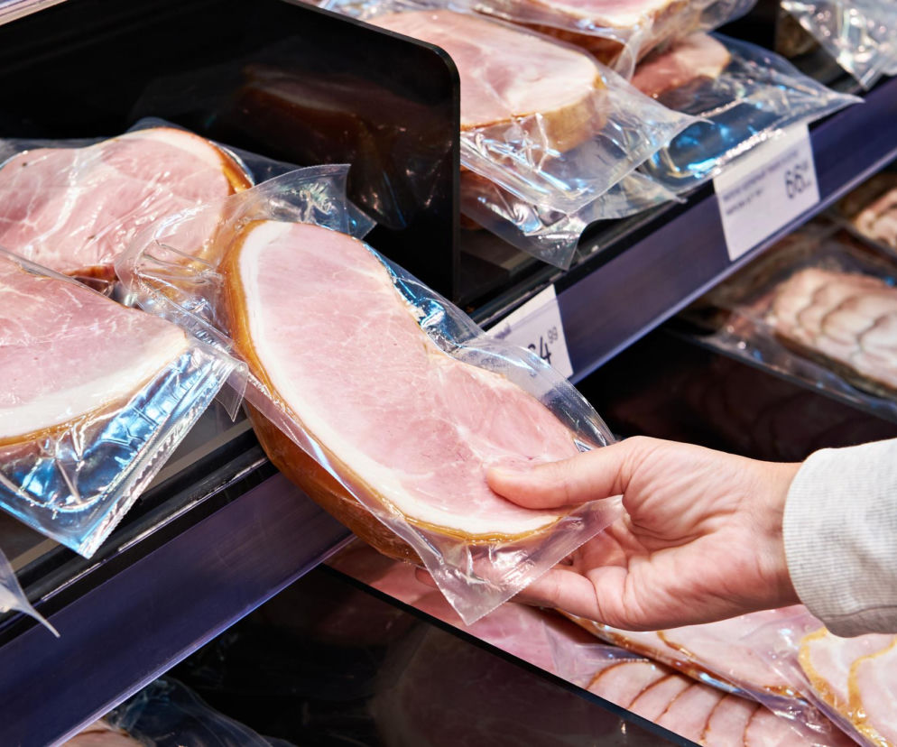 Dłoń osoby w białej bluzie sięgająca po hermetycznie zapakowaną wędlinę w plasterkach z półki sklepowej. Na opakowaniach widoczne etykiety cenowe. Obraz podkreśla temat świadomych wyborów żywieniowych i ryzyka związanego z przetworzoną żywnością, o czym przeczytasz więcej na Poradnik Zdrowie.